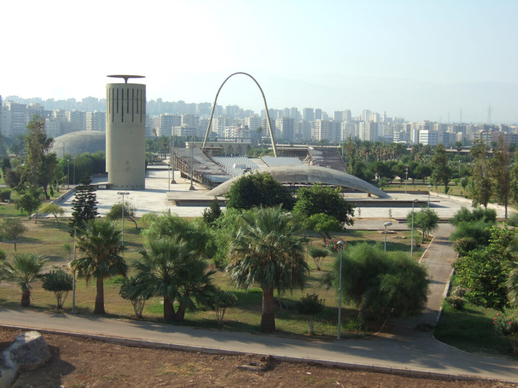 View of the Rachid Karami International Fair in Tripoli, Lebanon, showing modernist concrete structures set within landscaped grounds and the city skyline in the background.