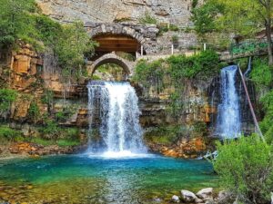 Waterfall cascading beneath historic stone arches into a turquoise pool surrounded by greenery and rock formations.