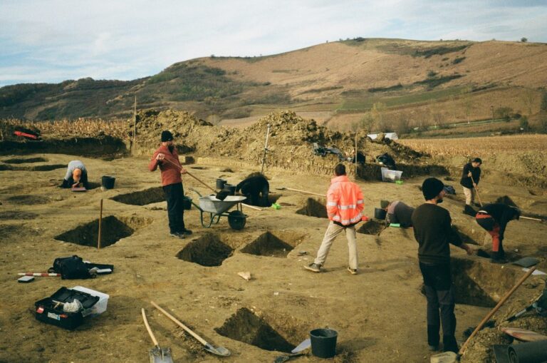 Archaeological excavation site with several team members working in open trenches across a rural landscape.