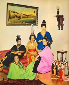 Five women wearing traditional Lebanese folkloric costumes pose indoors, seated and standing around a low seating area, with decorative objects and a framed landscape painting behind them.