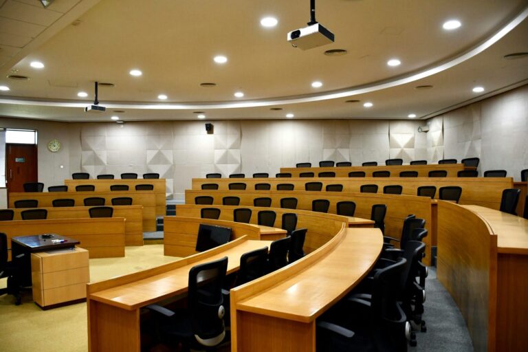 Modern lecture hall with tiered wooden seating, desks, and ceiling-mounted projectors in an academic setting.