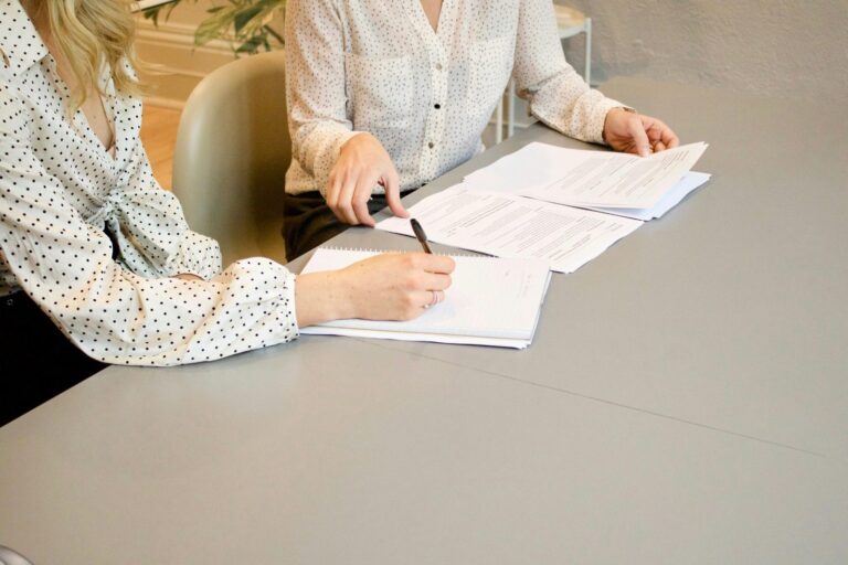 Two professionals reviewing documents and taking notes at a desk during a working session.